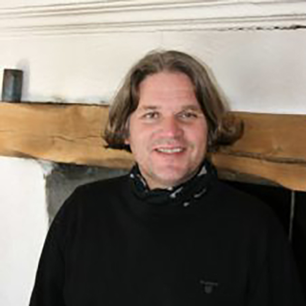 Mann med brunt hår med sort skjorte og halstørkle foran en peis.